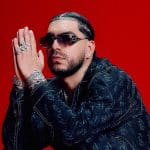 A man with a short hairstyle and facial hair poses against a red background, wearing a black patterned jacket over a dark shirt. he has multiple rings, a watch, and earrings, with his hands clasped near his face.