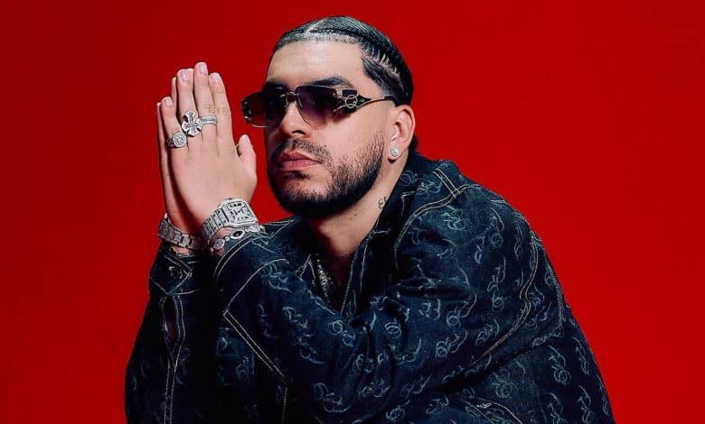 A man with a short hairstyle and facial hair poses against a red background, wearing a black patterned jacket over a dark shirt. he has multiple rings, a watch, and earrings, with his hands clasped near his face.