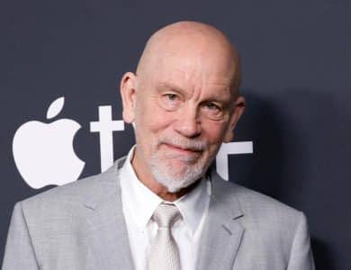 An older man with a bald head and white beard, wearing a light gray suit and tie, posing in front of a backdrop featuring the apple logo at an event.