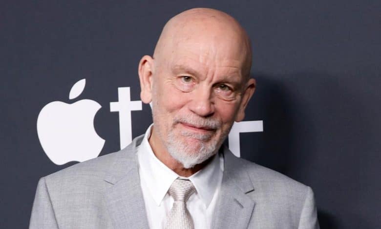 An older man with a bald head and white beard, wearing a light gray suit and tie, posing in front of a backdrop featuring the apple logo at an event.