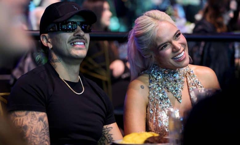 A man and a woman seated at a table, smiling and enjoying an event. the man wears a black cap and sunglasses, the woman in a sparkling silver dress. their expressions suggest they are having a good time.