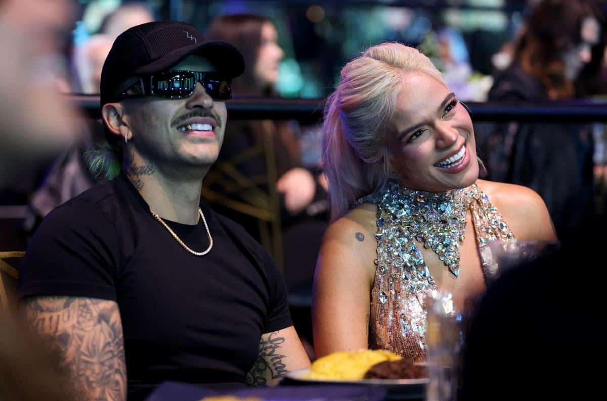 A man and a woman seated at a table, smiling and enjoying an event. the man wears a black cap and sunglasses, the woman in a sparkling silver dress. their expressions suggest they are having a good time.