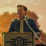 A middle-aged man in a blue astronaut suit with a golden "33" badge stands at a microphone, smiling confidently. he is outdoors, with trees and a blurred farm silo in the golden-hued background.