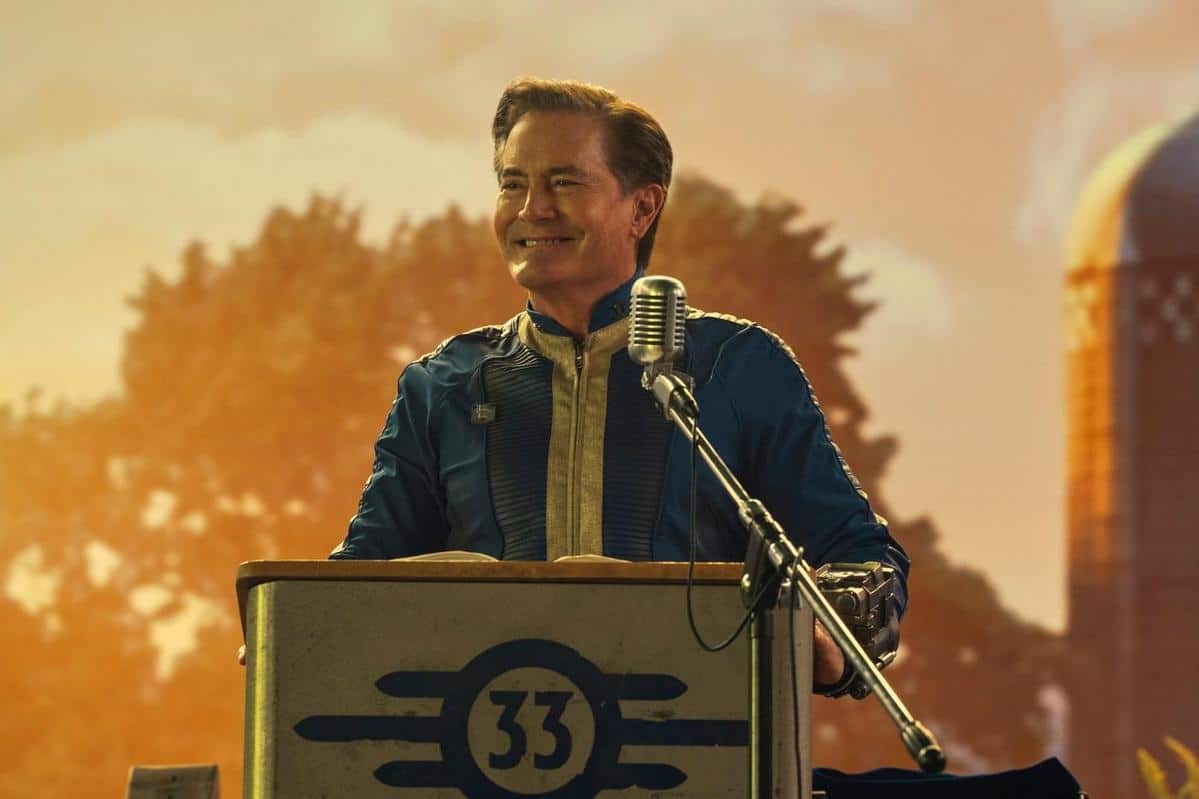 A middle-aged man in a blue astronaut suit with a golden "33" badge stands at a microphone, smiling confidently. he is outdoors, with trees and a blurred farm silo in the golden-hued background.