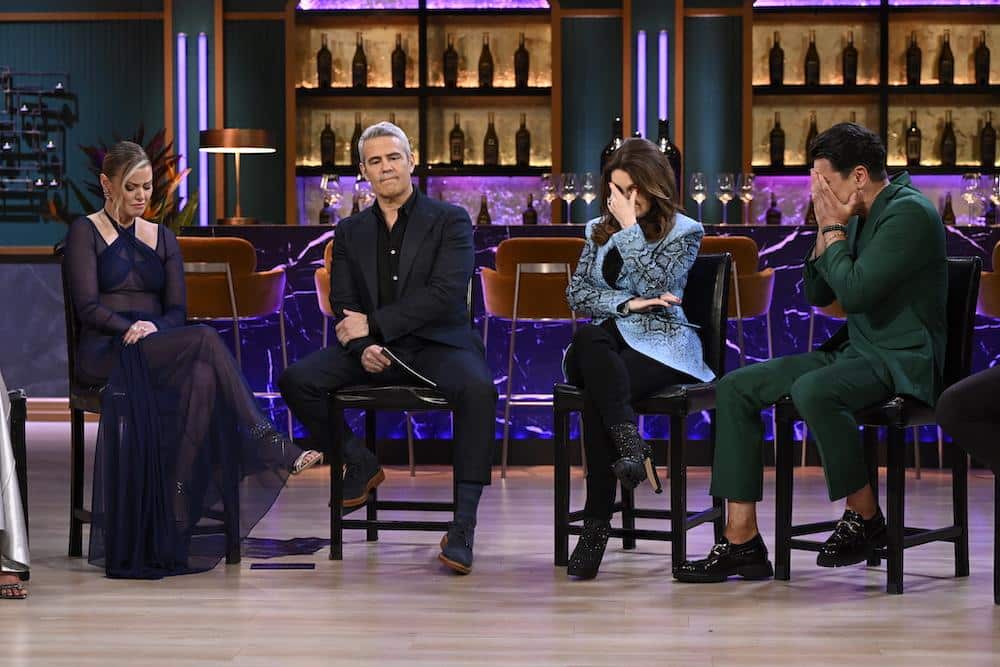 Four people seated on high stools on a tv set that resembles a bar, with shelves of bottles in the background. two women and two men appear engaged in a serious conversation. the setting is well-lit and modern.