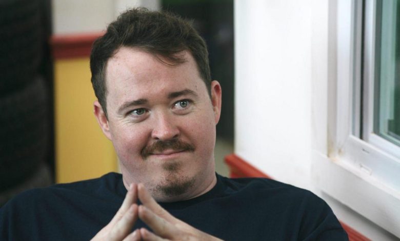 A man with a light complexion and short, dark hair smirks slightly. he wears a navy blue shirt and sits with his fingers interlocked, resting under his chin. he's beside a window with red and white frames, suggesting a casual setting.