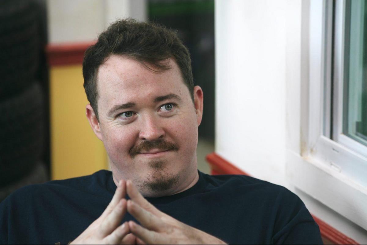 A man with a light complexion and short, dark hair smirks slightly. he wears a navy blue shirt and sits with his fingers interlocked, resting under his chin. he's beside a window with red and white frames, suggesting a casual setting.