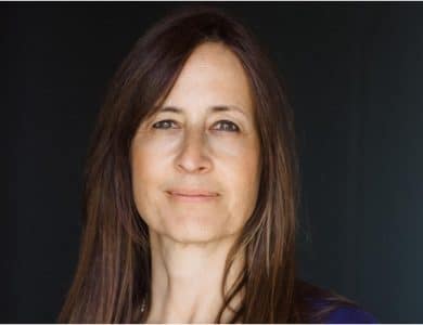 A portrait of a middle-aged woman with long, straight brown hair and a gentle smile. she wears a blue top and stands before a dark background, facing the camera with a calm, welcoming expression.