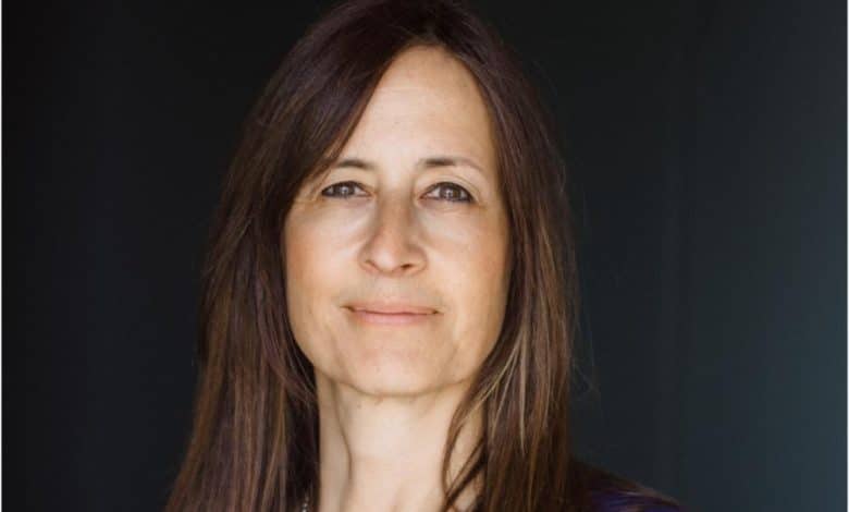 A portrait of a middle-aged woman with long, straight brown hair and a gentle smile. she wears a blue top and stands before a dark background, facing the camera with a calm, welcoming expression.