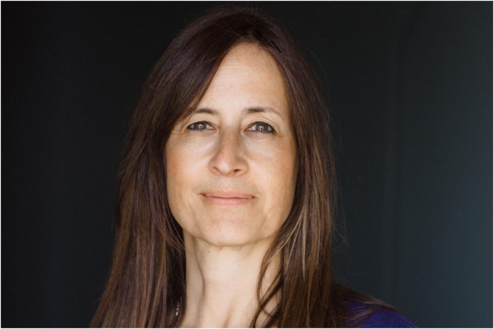 A portrait of a middle-aged woman with long, straight brown hair and a gentle smile. she wears a blue top and stands before a dark background, facing the camera with a calm, welcoming expression.