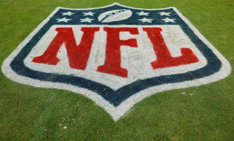 The image shows the nfl logo painted on green grass. the logo features the letters "nfl" in red, and a blue outline of a football above a set of stars, with a blue border.