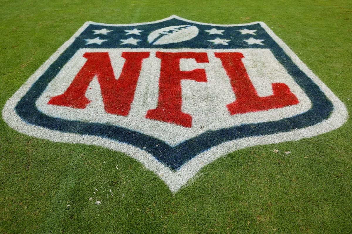 The image shows the nfl logo painted on green grass. the logo features the letters "nfl" in red, and a blue outline of a football above a set of stars, with a blue border.