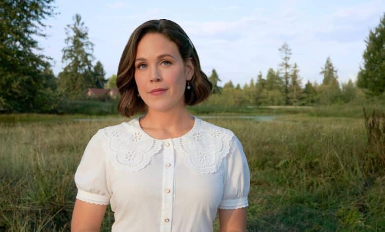 A woman with medium-length brunette hair stands outdoors, wearing a white blouse with lace detailing. she gazes directly at the camera with a calm expression, set against a backdrop of a serene landscape with trees and a field.