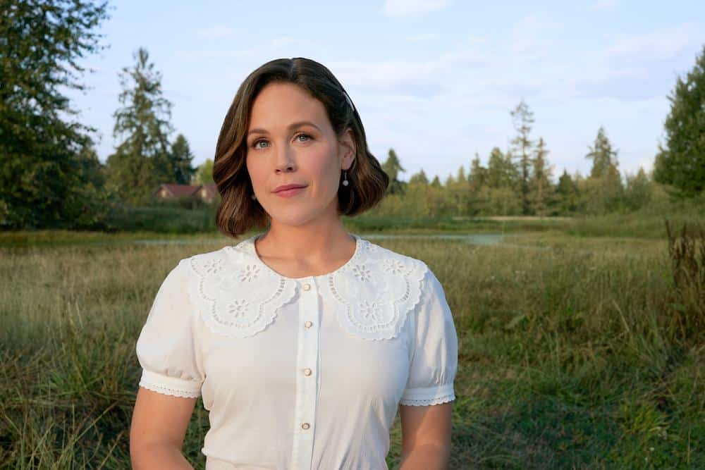 A woman with medium-length brunette hair stands outdoors, wearing a white blouse with lace detailing. she gazes directly at the camera with a calm expression, set against a backdrop of a serene landscape with trees and a field.