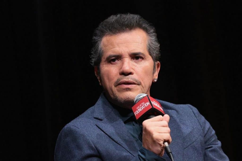 A man with short, dark hair and a trimmed goatee wears a dark blue blazer and black shirt. He holds a microphone labeled “FOUNDATION” and appears to be speaking on stage, looking slightly to the side. The background is dark.