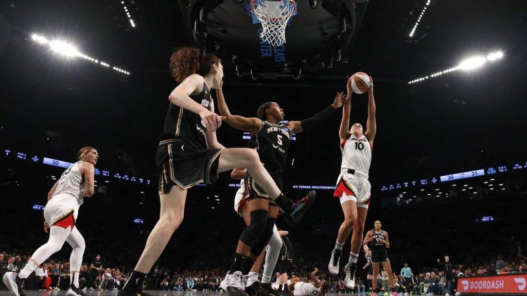 A women’s basketball player in a white uniform leaps to shoot the ball near the hoop, while three defenders in black try to block her. The action takes place in a packed arena, with the crowd and scoreboard visible in the background.