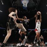 A women’s basketball player in a white uniform leaps to shoot the ball near the hoop, while three defenders in black try to block her. The action takes place in a packed arena, with the crowd and scoreboard visible in the background.