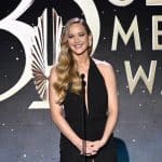 A woman with long, wavy blonde hair wearing a black halter dress stands smiling at a microphone on stage. Behind her, a large illuminated sign reads “30th CLA MEDIA AWARDS” in elegant gold letters on a dark, starry background.
