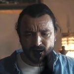 A man with dark hair and a beard looks down with a distressed expression, wearing a light blue shirt over a white tee. Black liquid drips from his mouth. The background is dimly lit with a vintage lamp and a framed picture on the wall.