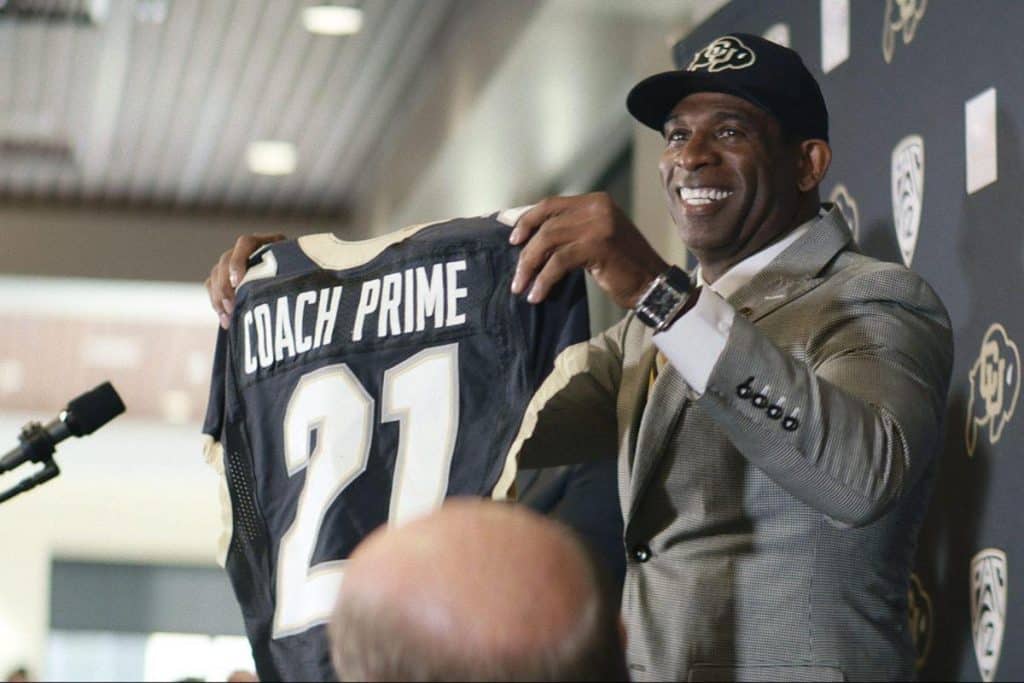 A smiling man in a suit and black cap holds up a black football jersey with "COACH PRIME" and the number 21. He stands at a podium with microphones, in front of a backdrop featuring the Colorado Buffaloes and PAC-12 logos.