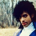 A person with curly black hair and bold eyeliner wears a royal blue jacket and white ruffled shirt, looking directly at the camera with a serious expression. The background shows bare trees and a muted, outdoor winter or autumn scene.