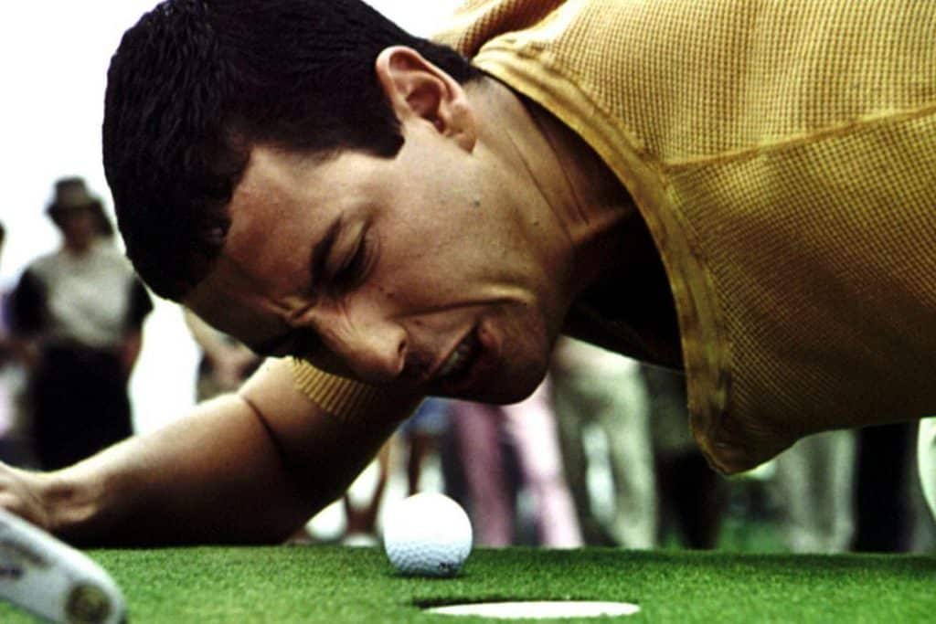 A man in a yellow shirt is lying close to the ground on a golf course, grimacing and intensely staring at a golf ball near the hole, with his face very close to the ball and the putter visible beside him. Blurry spectators are in the background.