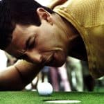 A man in a yellow shirt is lying close to the ground on a golf course, grimacing and intensely staring at a golf ball near the hole, with his face very close to the ball and the putter visible beside him. Blurry spectators are in the background.