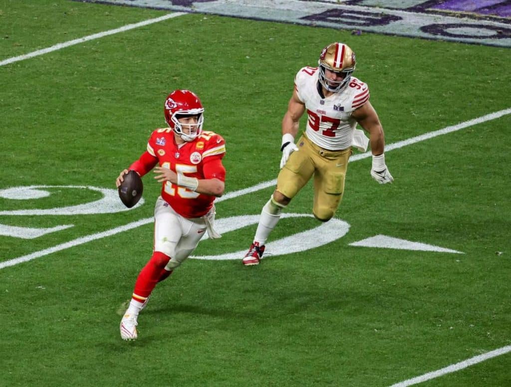 A football player in a red Kansas City Chiefs uniform runs with the ball, looking downfield to pass, as a San Francisco 49ers player in a white and gold uniform chases him during a game on a green field with white yard lines and marks visible.