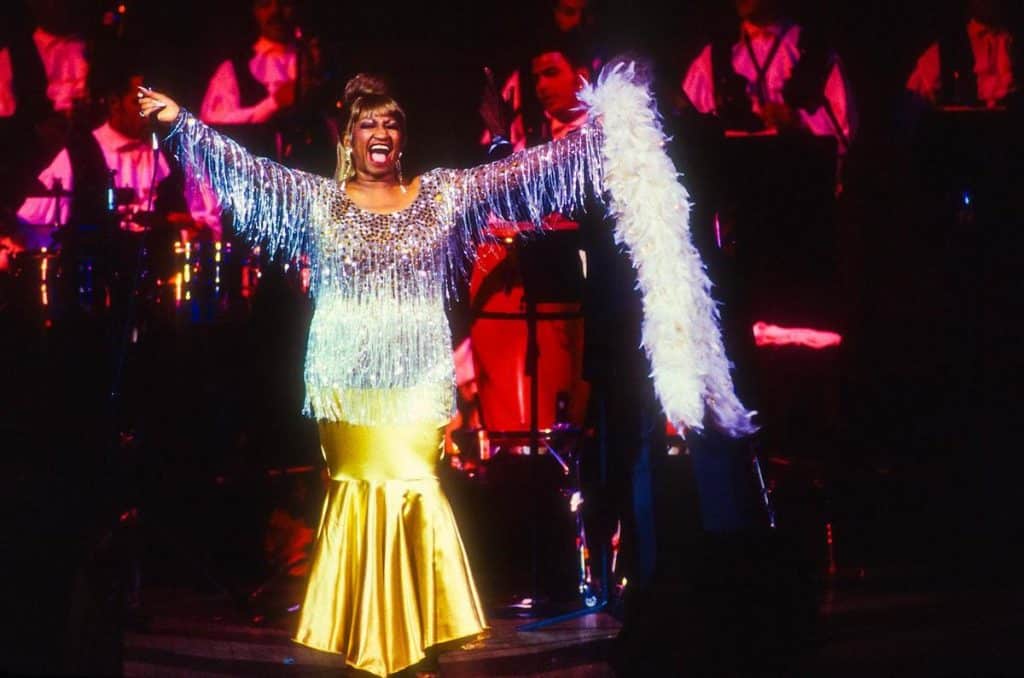 A singer in a sparkling, fringed silver top and gold skirt performs on stage with arms outstretched. She is smiling widely, wearing a white feather boa, and is illuminated by colorful stage lights, with a band playing in the background.