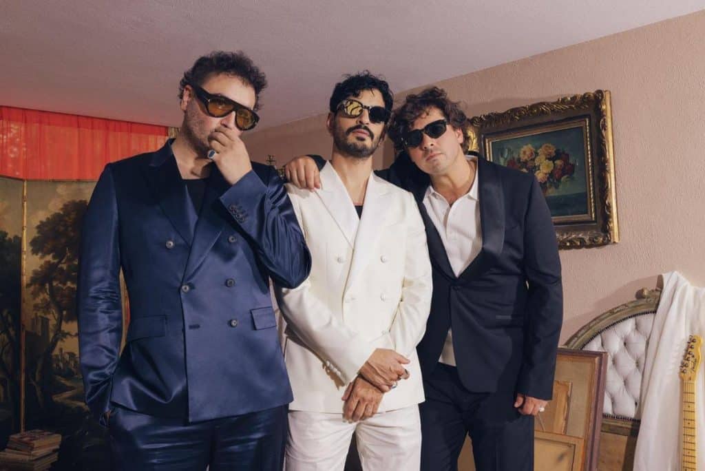 Three men wearing suits and sunglasses stand indoors. The man on the left wears a navy suit, the center man wears a white suit, and the right man wears a black suit. They pose confidently in a warmly lit room with paintings and vintage decor in the background.