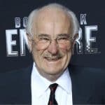 An older man with thinning gray hair, glasses, and a mustache smiles while wearing a black suit, white shirt, and black tie against a dark background with partially visible text reading "Boardwalk Empire.