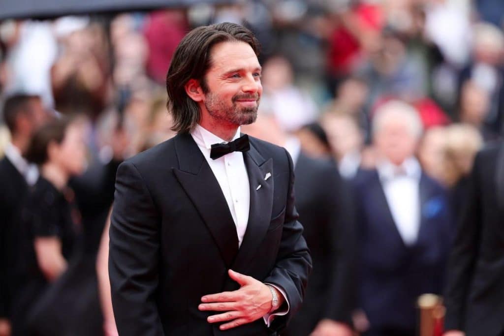 A man with medium-length dark hair and a beard, dressed in a black tuxedo with a black bow tie and white shirt, stands smiling on a red carpet at a formal event. Blurred crowds and photographers are visible in the background.
