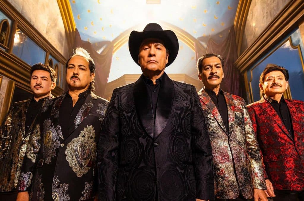 Five men stand facing forward in ornate, patterned suits. The man in the center wears a black cowboy hat and a black floral suit. The background features golden light and a blue, starry ceiling, resembling an elaborate indoor setting.