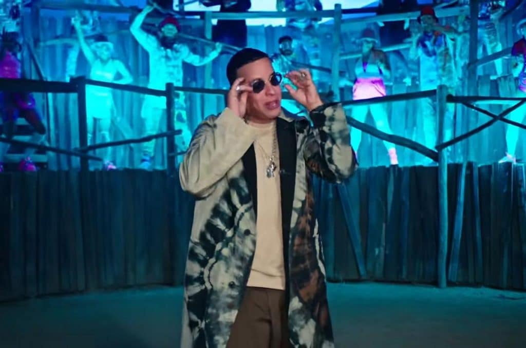 A man wearing sunglasses, a tie-dye coat, and light shirt stands in front of a wooden fence. Blue lighting illuminates him and a group of people dancing in the background, who appear frozen in energetic poses. The scene has a vibrant, party-like atmosphere.