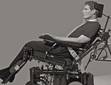 Black and white photo of a man using a complex, motorized wheelchair, equipped with various supports and devices. he wears dark clothing and has a focused expression, sitting against a plain background.