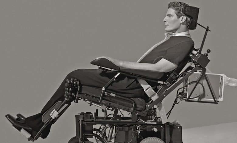 Black and white photo of a man using a complex, motorized wheelchair, equipped with various supports and devices. he wears dark clothing and has a focused expression, sitting against a plain background.