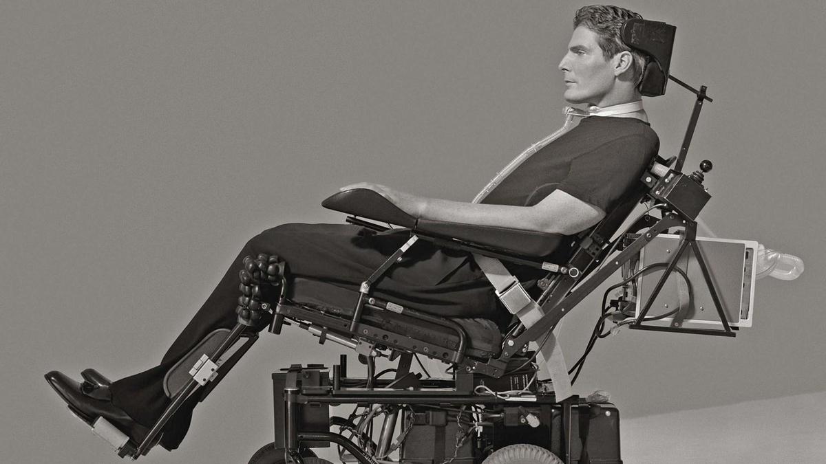 Black and white photo of a man using a complex, motorized wheelchair, equipped with various supports and devices. he wears dark clothing and has a focused expression, sitting against a plain background.