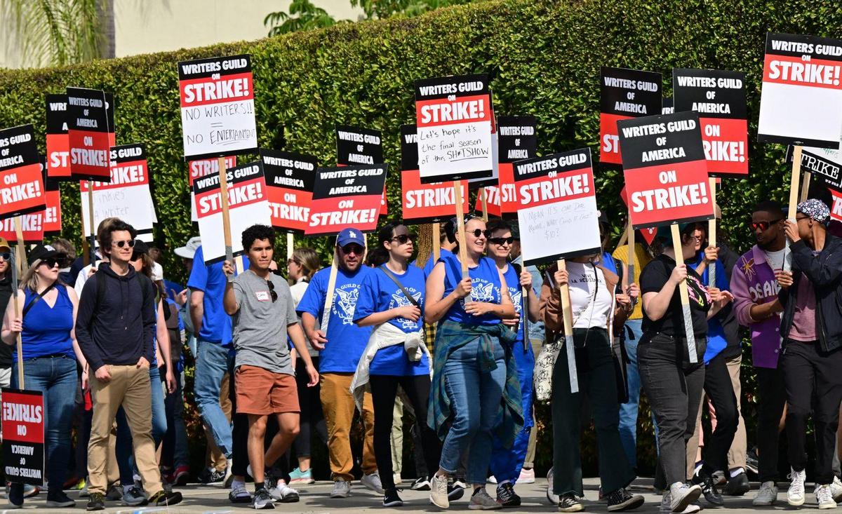 A group of people holding "writers guild strike" signs on a sunny day during a protest. they appear determined and united, and wear casual clothing as they march along a street lined with trees.