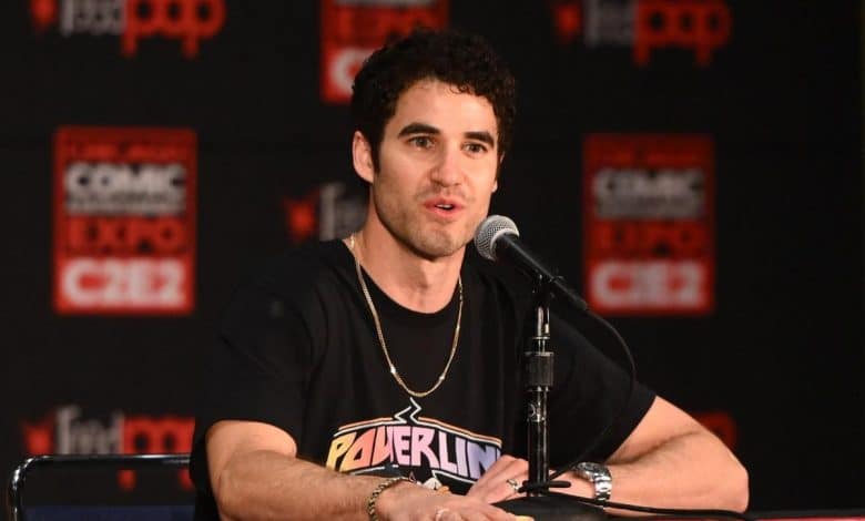 A man with curly dark hair and a stubble is sitting at a table speaking into a microphone at a comic expo event. he wears a black powerline t-shirt and is surrounded by a red backdrop with event logos.