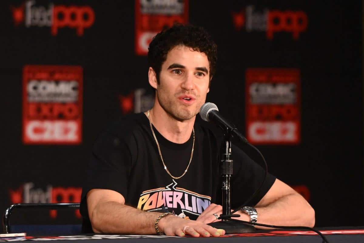 A man with curly dark hair and a stubble is sitting at a table speaking into a microphone at a comic expo event. he wears a black powerline t-shirt and is surrounded by a red backdrop with event logos.