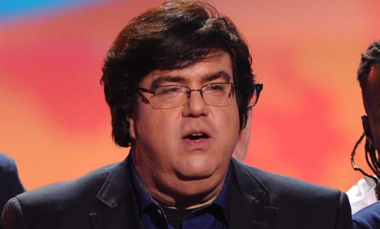 A man with tousled dark hair and wearing glasses speaks into a microphone at an event. he wears a navy suit over a black shirt. the background shows a gradient of red to blue light.