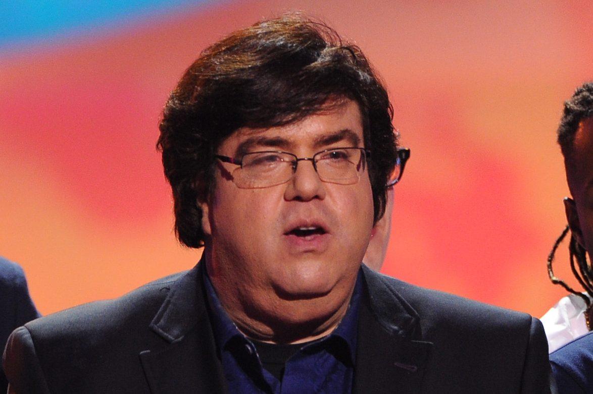 A man with tousled dark hair and wearing glasses speaks into a microphone at an event. he wears a navy suit over a black shirt. the background shows a gradient of red to blue light.