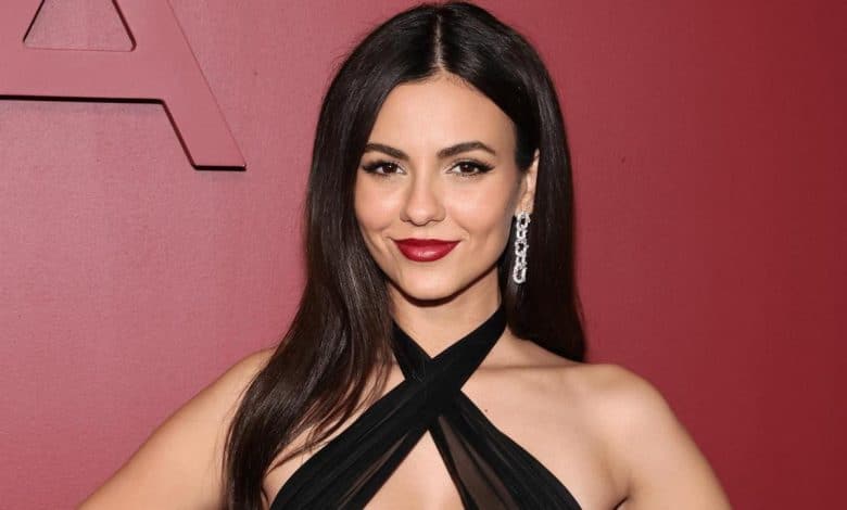 A woman with long dark hair, wearing a stylish black halter neck dress and long silver earrings, poses confidently against a maroon background. her makeup is elegant, featuring bold red lipstick and defined eyebrows.