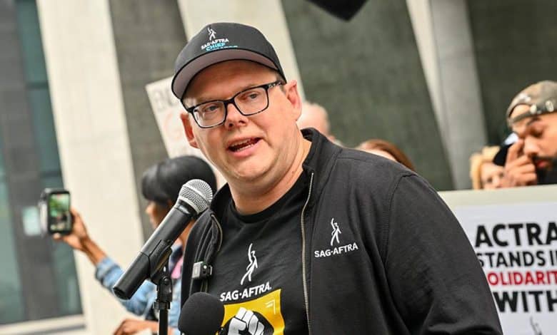 A man wearing glasses speaks into a microphone at an outdoor event, donning a black cap and t-shirt with sag-aftra logos. behind him, participants hold signs, visibly engaged. there's a building backdrop.