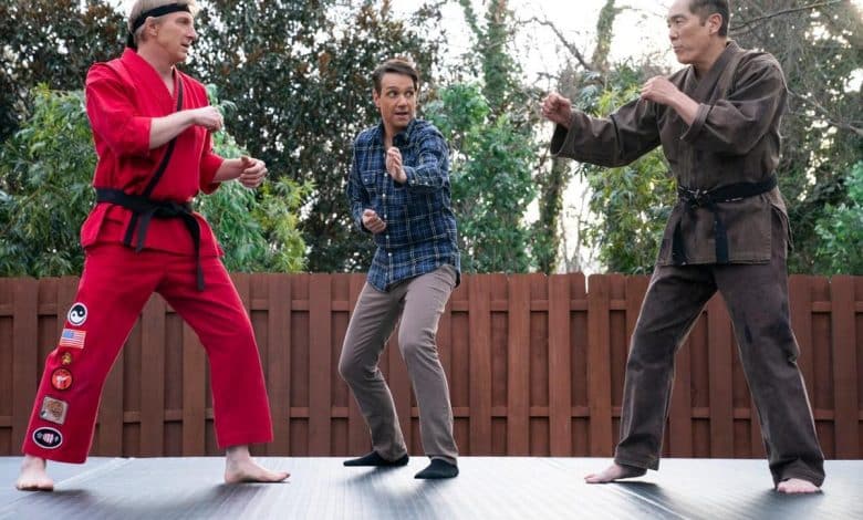 Three men on a dojo mat outdoors; one in a red karate gi, one in casual clothing looking surprised, and one in a brown traditional martial arts uniform, all in defensive stances.
