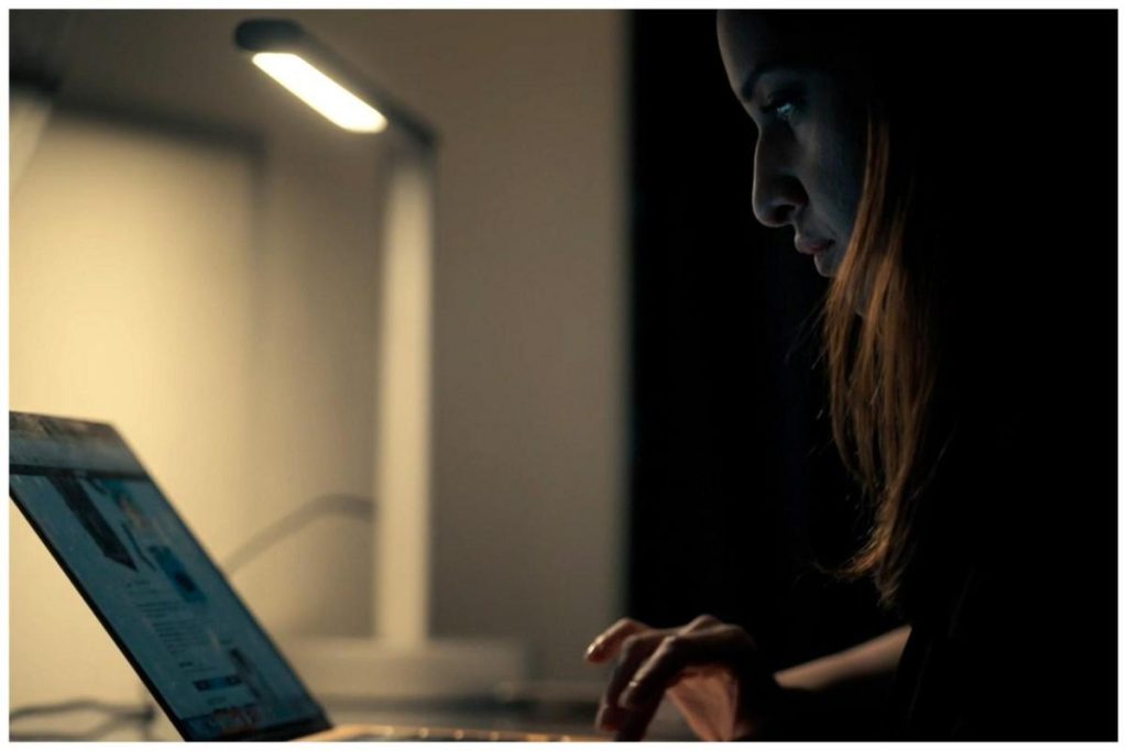 A woman focuses intently on her laptop screen under a desk lamp in a dimly lit room, highlighting her profile and the glow from the screen.