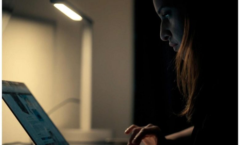 A woman focuses intently on her laptop screen under a desk lamp in a dimly lit room, highlighting her profile and the glow from the screen.