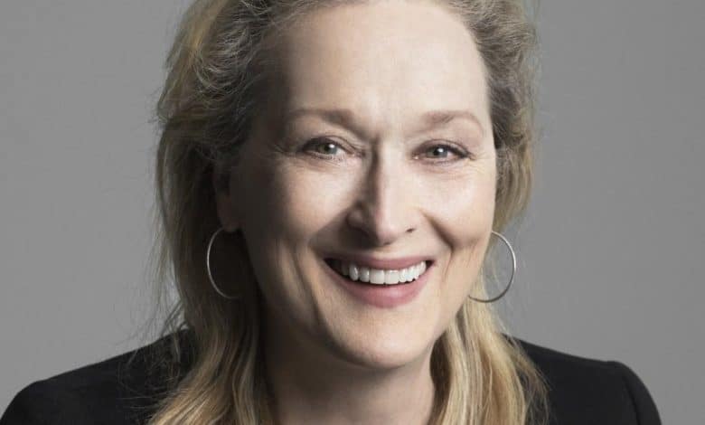 A close-up portrait of a smiling caucasian woman with long, light blonde hair. she wears large hoop earrings and a black blazer. her expression is joyful and welcoming, set against a gray background.