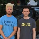 Two men stand in front of a blurred car and floral background. the man on the left has a blond buzz cut and wears a blue shirt labeled "party rock," and the man on the right, sporting short dark hair, wears a black shirt with "skull" printed on it.