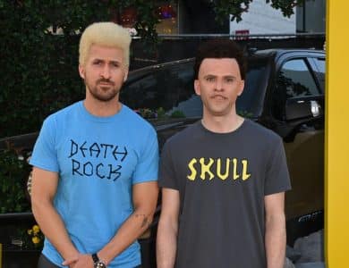 Two men stand in front of a blurred car and floral background. the man on the left has a blond buzz cut and wears a blue shirt labeled "party rock," and the man on the right, sporting short dark hair, wears a black shirt with "skull" printed on it.